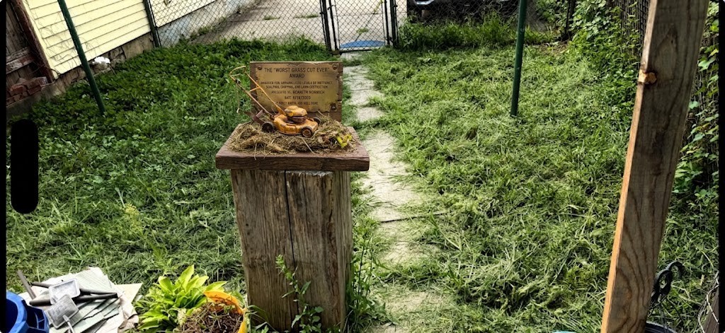 The Worst Grass Cut Ever award displayed on a wooden pedestal in the yard where the grass was cut.